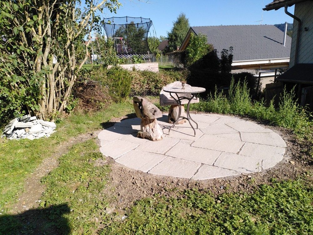 Terrasse im Hinterhof mit runden Steinplatten, einem kleinen Tisch und Stühlen, umgeben von Grün