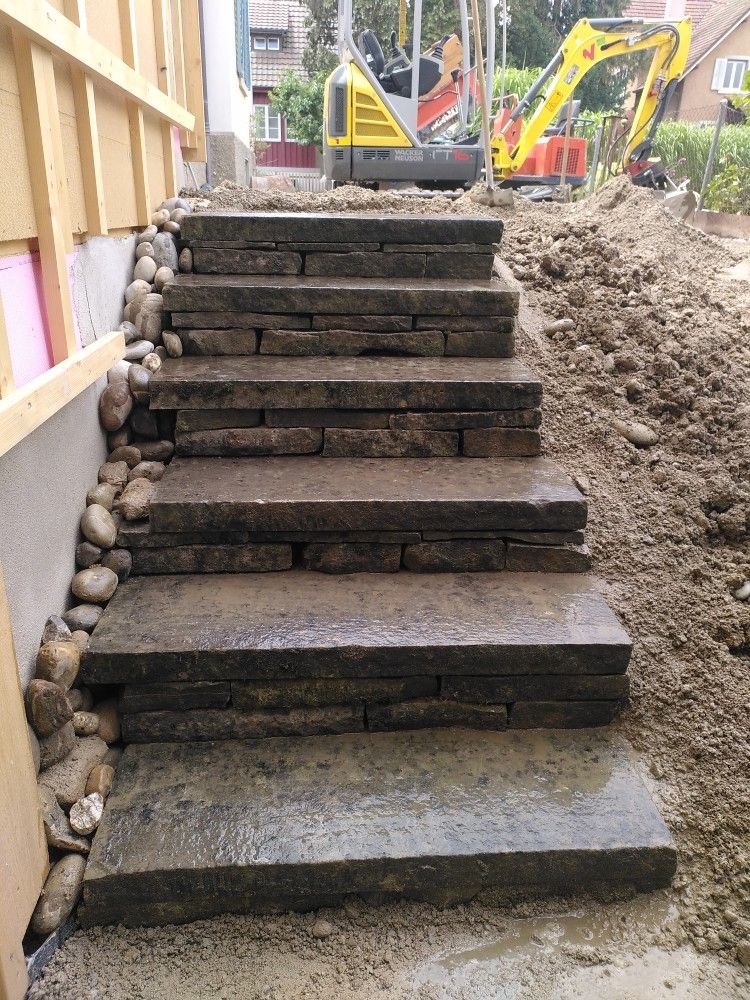 Neben einem Haus werden Steintreppen gebaut, im Hintergrund sind Bagger und Erde zu sehen.