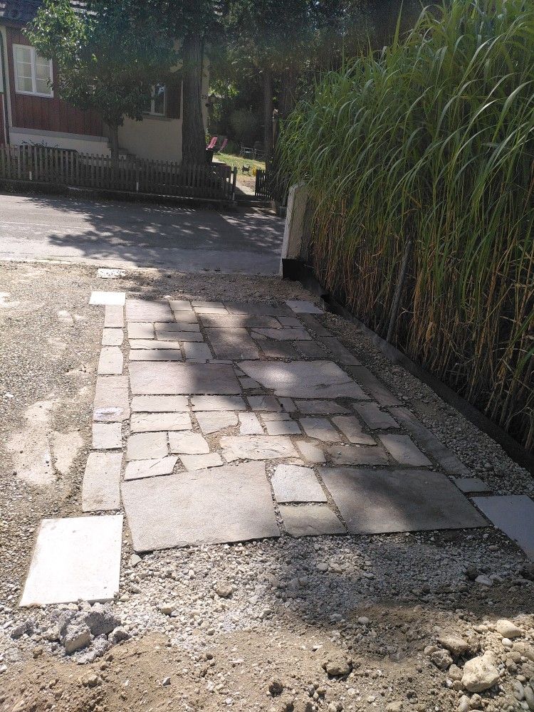 Ein Gehweg aus zerklüfteten Steinen neben einem grasbewachsenen Zaun, mit einem rauen Kiesrand und hellem Sonnenlicht.
