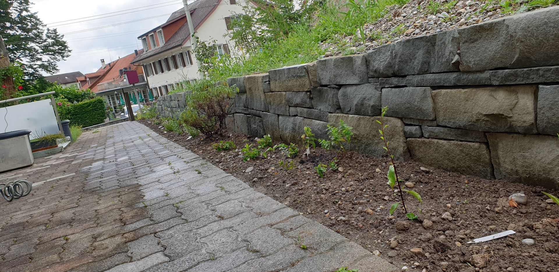 Ein mit Steinen gepflasterter Weg verläuft neben einer niedrigen Steinmauer; in der Ferne sind Pflanzen und Häuser zu sehen.
