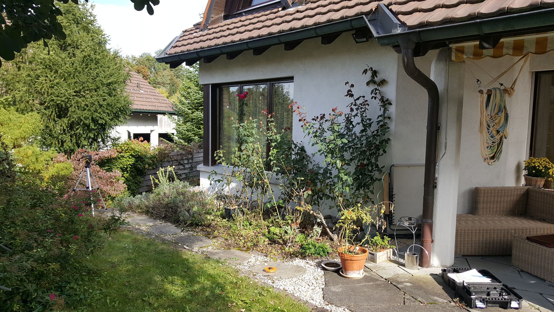 Gartenterrasse neben einem Haus mit Topfpflanzen, Glasschiebetüren und einem Korbsessel unter einem Ziegeldach.