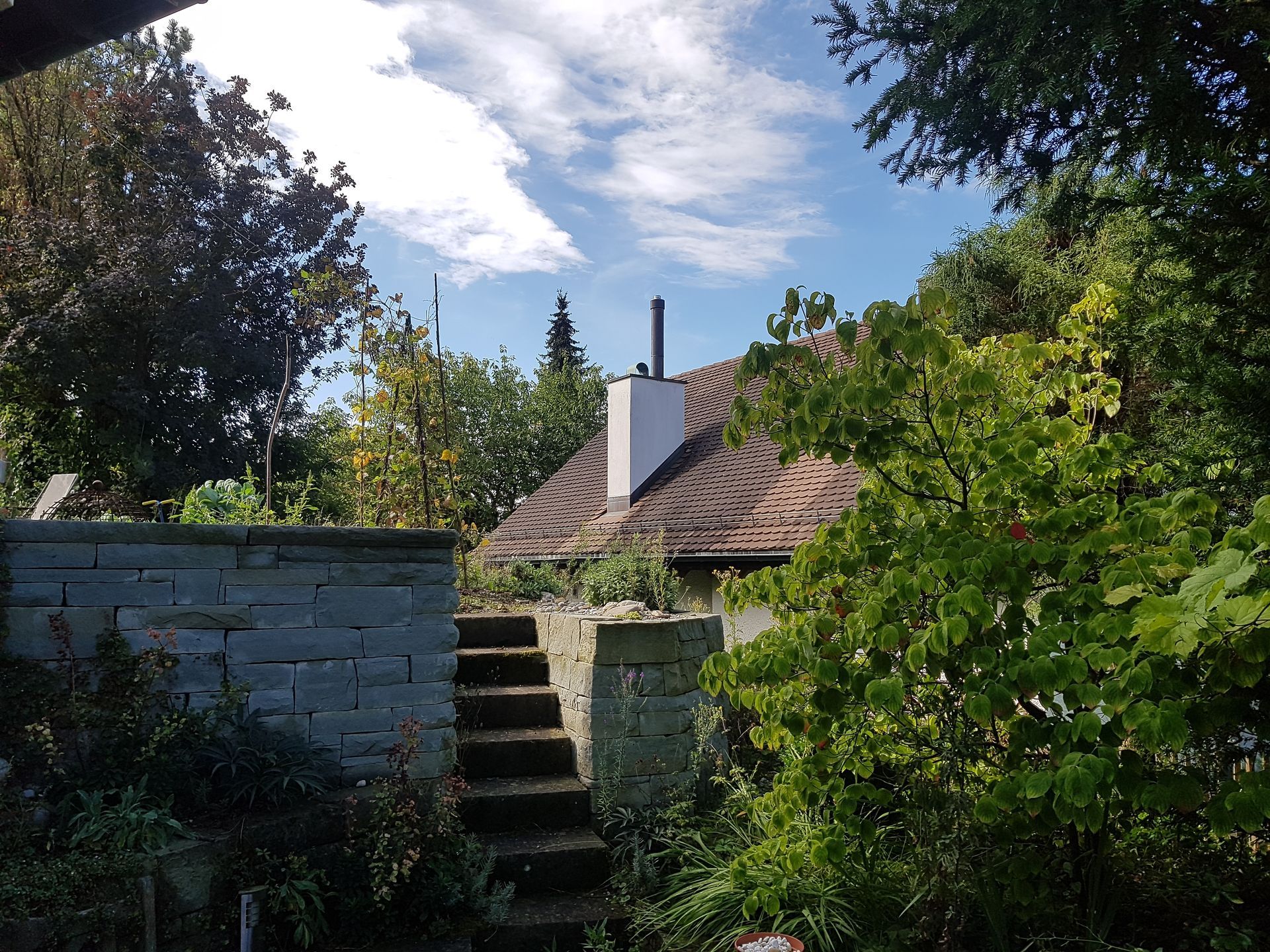 Eine Gartentreppe führt zu einem Haus mit Ziegeldach inmitten üppigen Grüns unter einem teilweise bewölkten Himmel