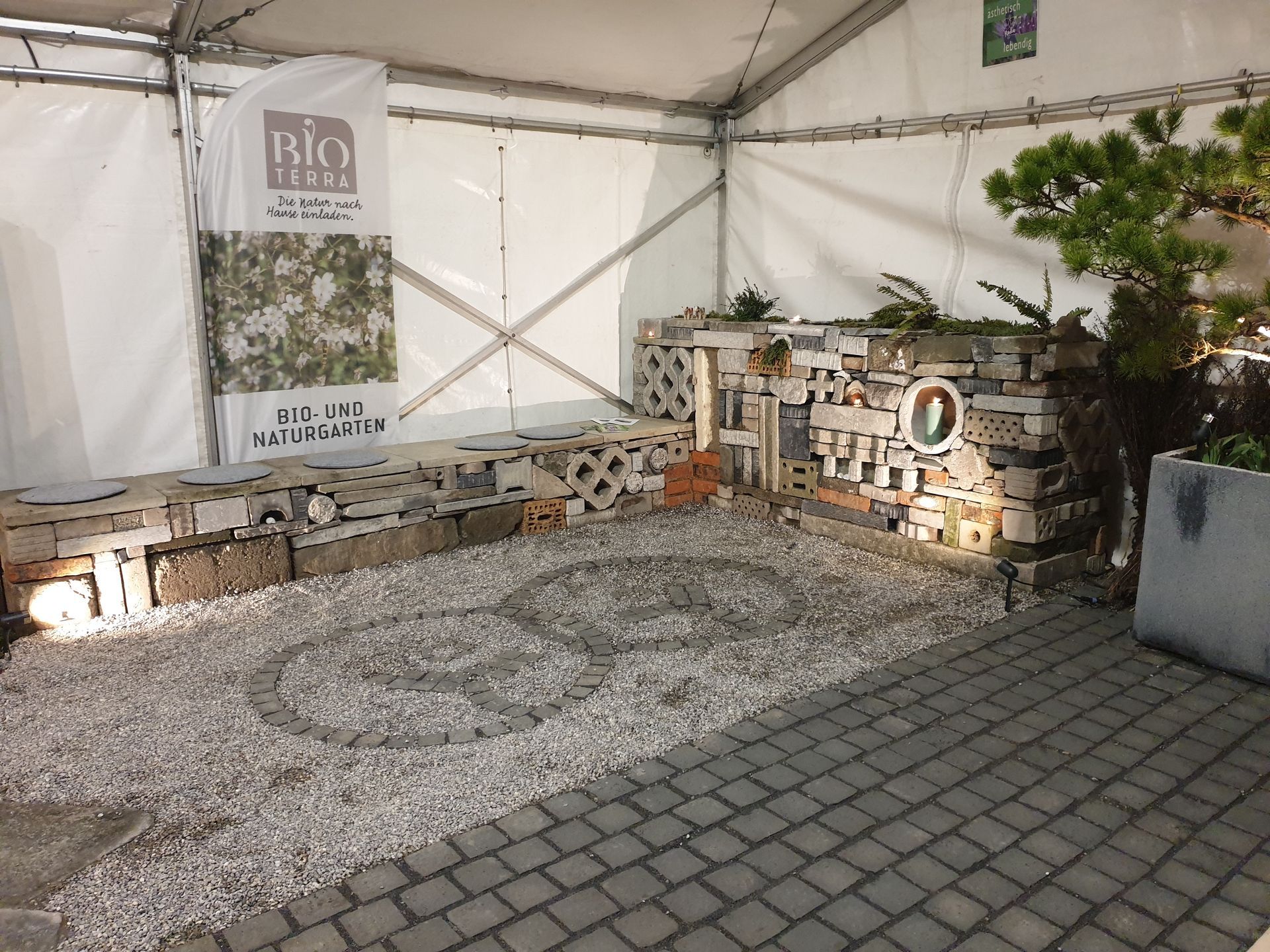 Weisses Zelt als Ausstellungsfläche mit Kiessitzen, Steinbänken und einem Schild unter einem Holzdach.