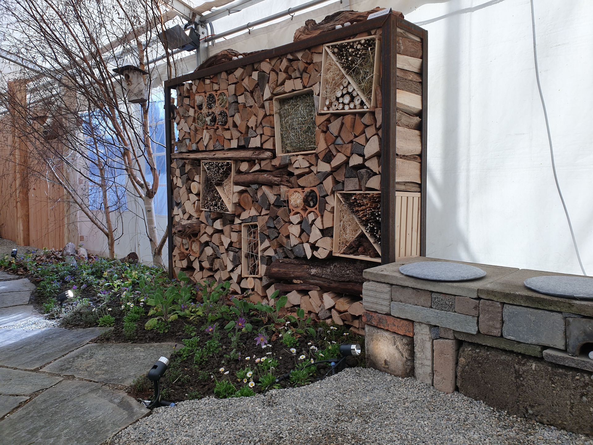 Ein Holzregal mit gespaltenem Brennholz neben einer Gartenmauer und einem Pflanzbeet