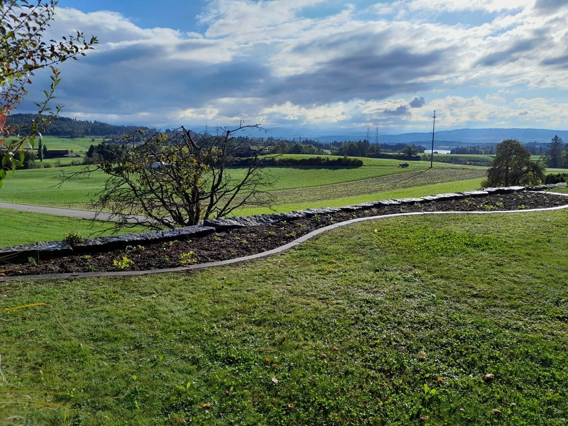 Grasbewachsener Park mit Blick auf ein Tal unter bewölktem Himmel, im Vordergrund eine Steinmauer