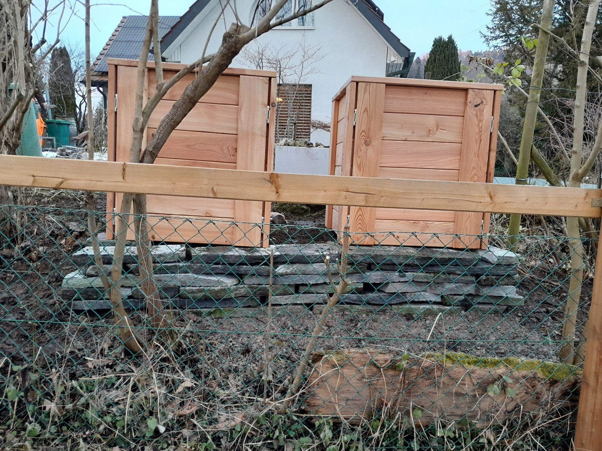 Zwei Holzkisten auf einer Steinmauer in einem Hof, dahinter ein Haus und kahle Äste.