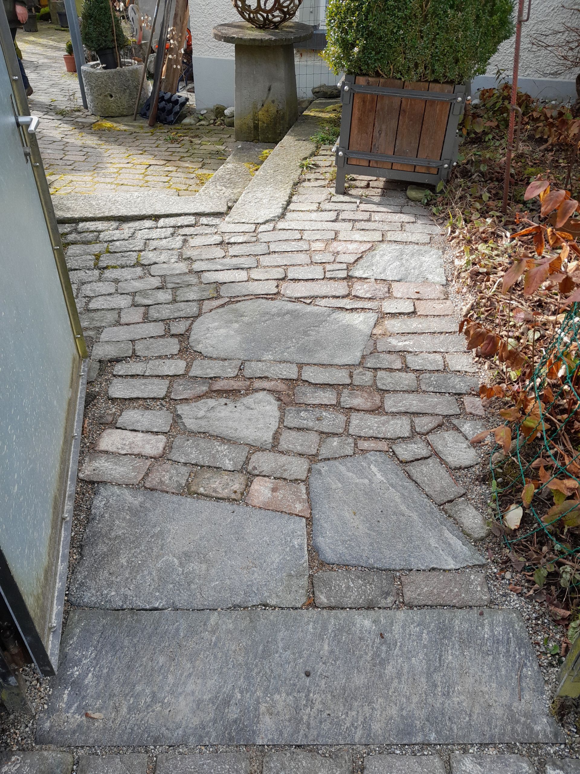 Schmaler Gartenweg mit unebenem Steinpflaster und Kopfsteinpflaster neben Sträuchern und einem Pflanzkasten
