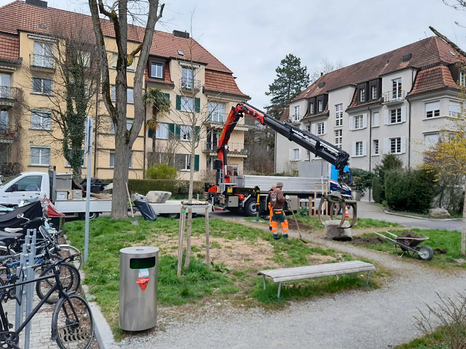 Ein Baukran hebt Baumaterialien in einem Innenhof neben Wohnhäusern und Fahrrädern an.