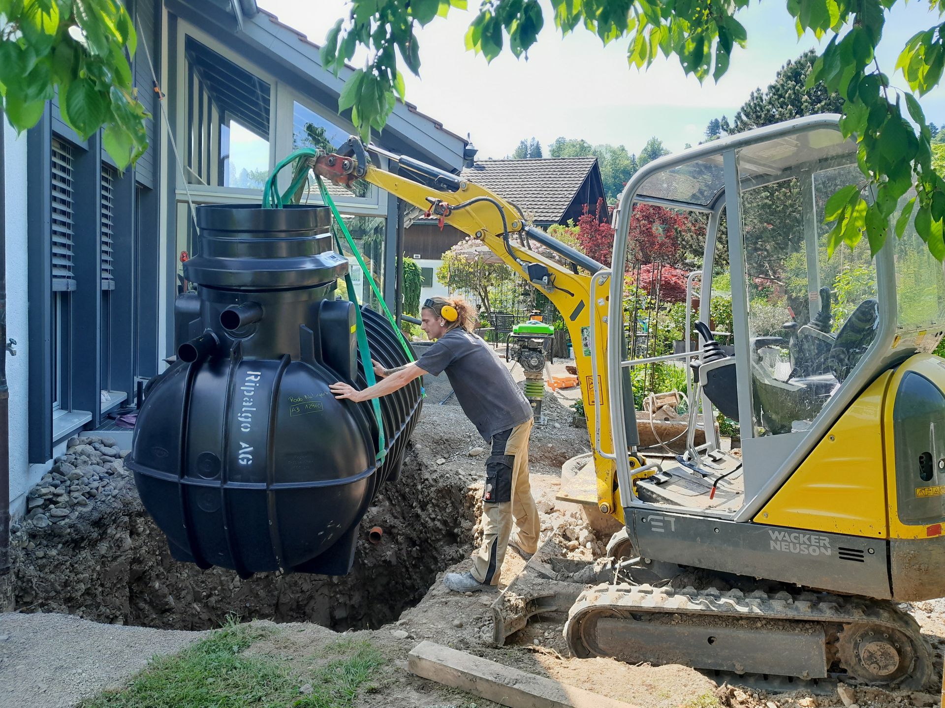 Ein Arbeiter installiert mit einem gelben Minibagger einen grossen schwarzen unterirdischen Tank neben einem Haus.