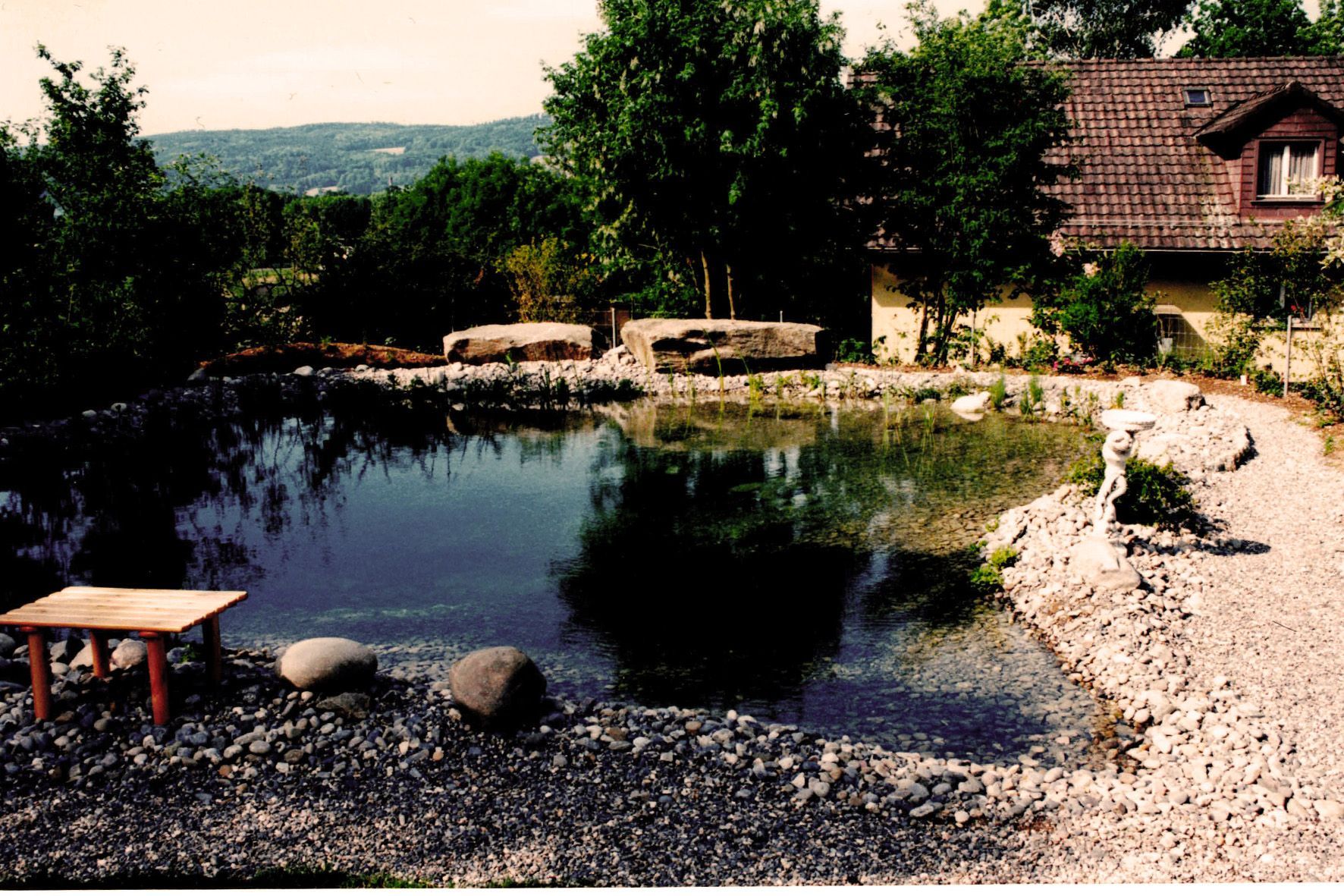 Kleiner Teich mit Steinrand, Kiesufer, Bäumen und einem rustikalen Haus im Hintergrund