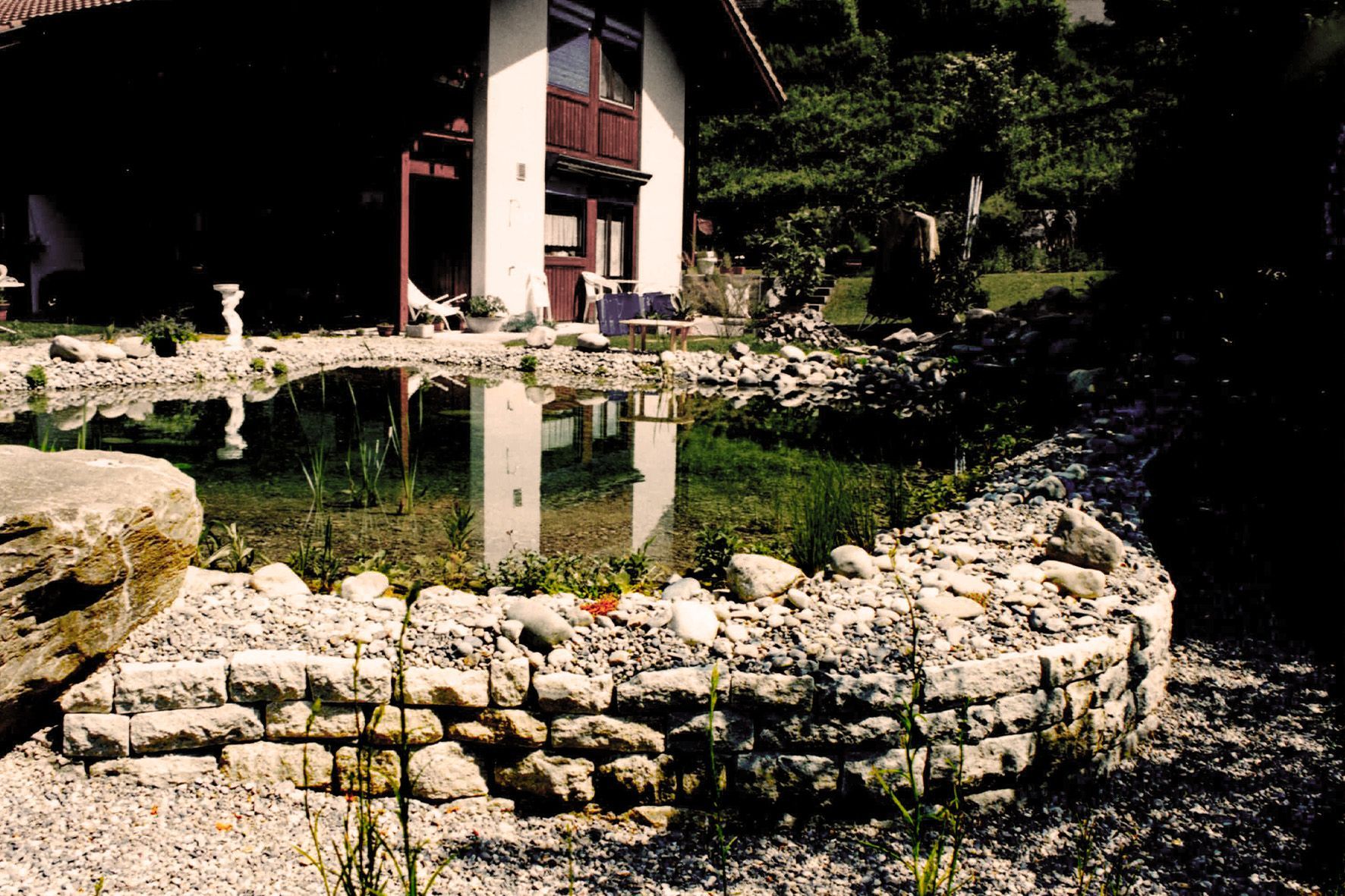 Ein Steinteich neben einem Gebäude mit rotem Dach spiegelt sich im ruhigen Wasser in einer Gartenlandschaft.