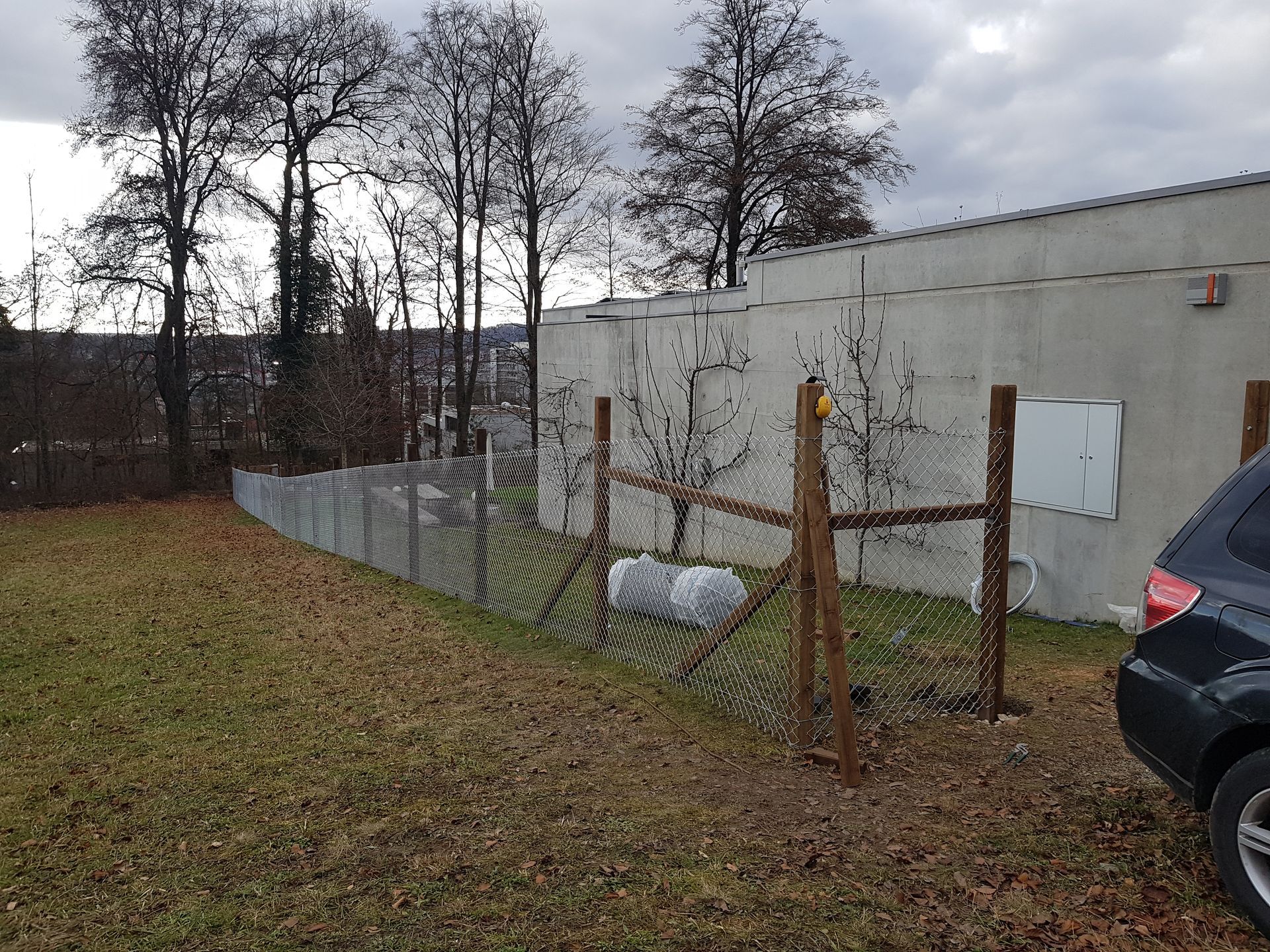 Ein mit Laub bedeckter Hof neben einem Maschendrahtzaun und einem weissen Gebäude, rechts davon ein geparktes Auto.