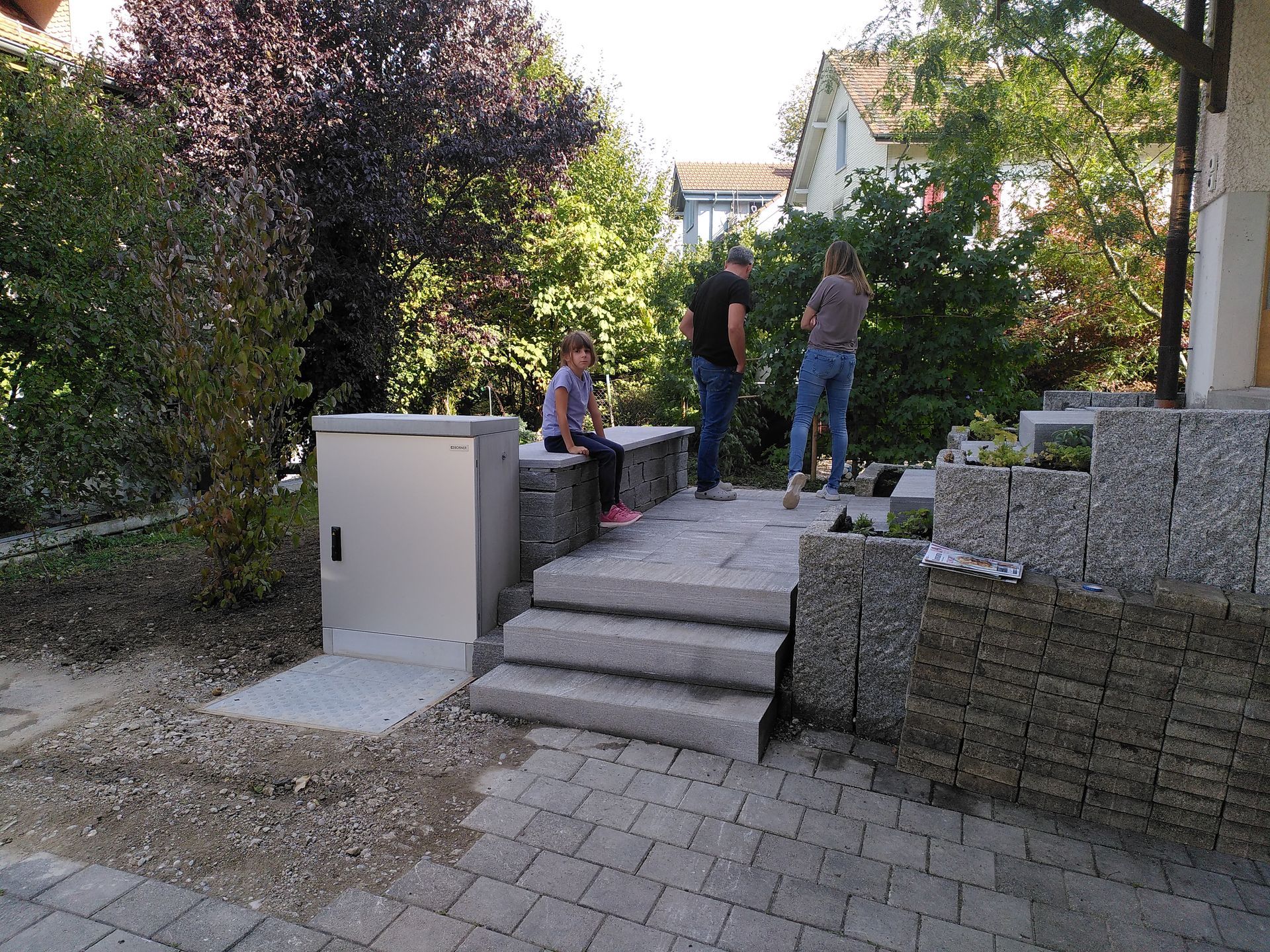 Menschen stehen auf Stufen in einer Gartenterrasse neben Steinblöcken und Grünpflanzen.