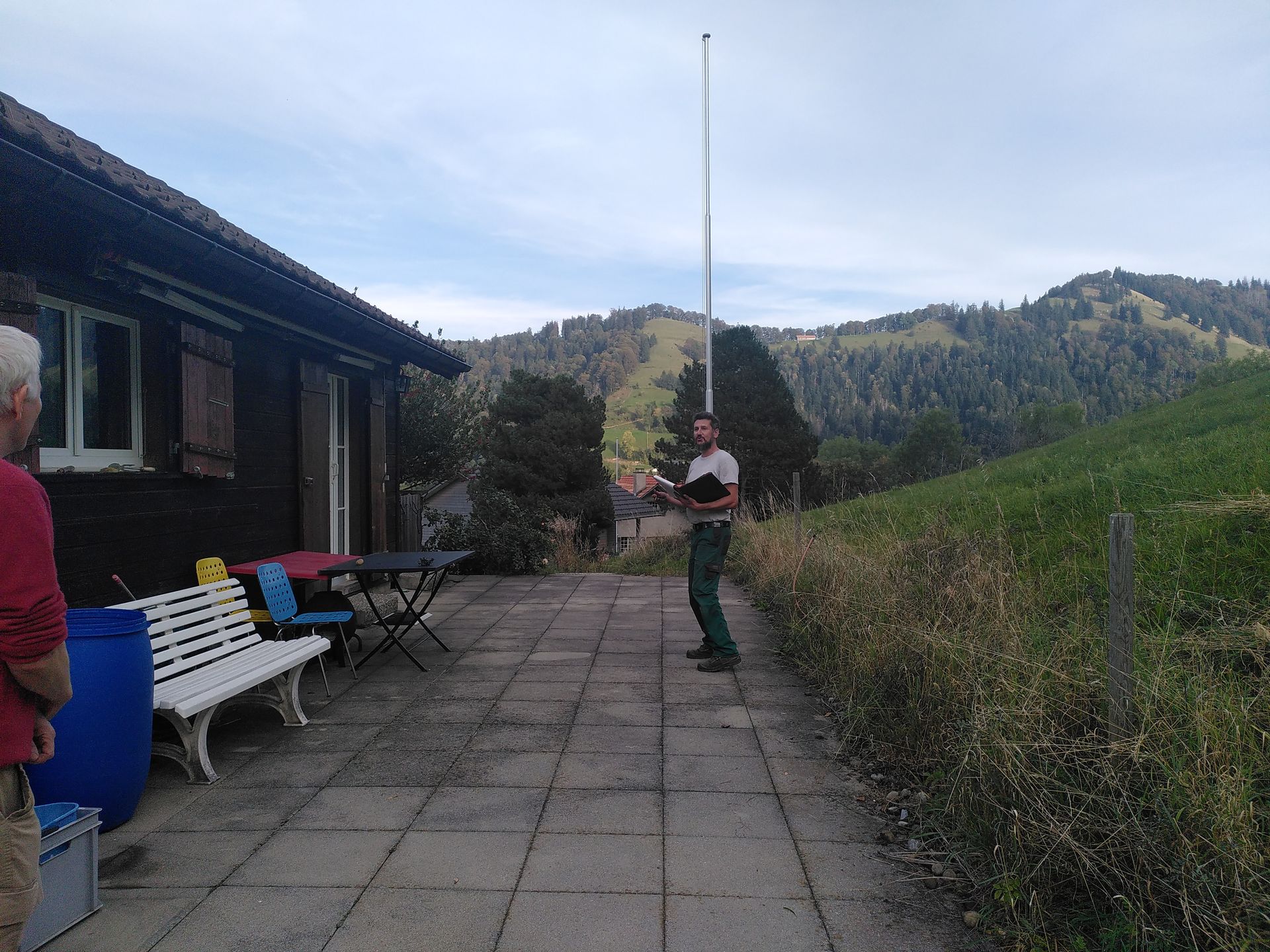 Eine Person steht auf einer Terrasse neben einem Gebäude und hält eine lange Stange; im Hintergrund erheben sich Hügel.