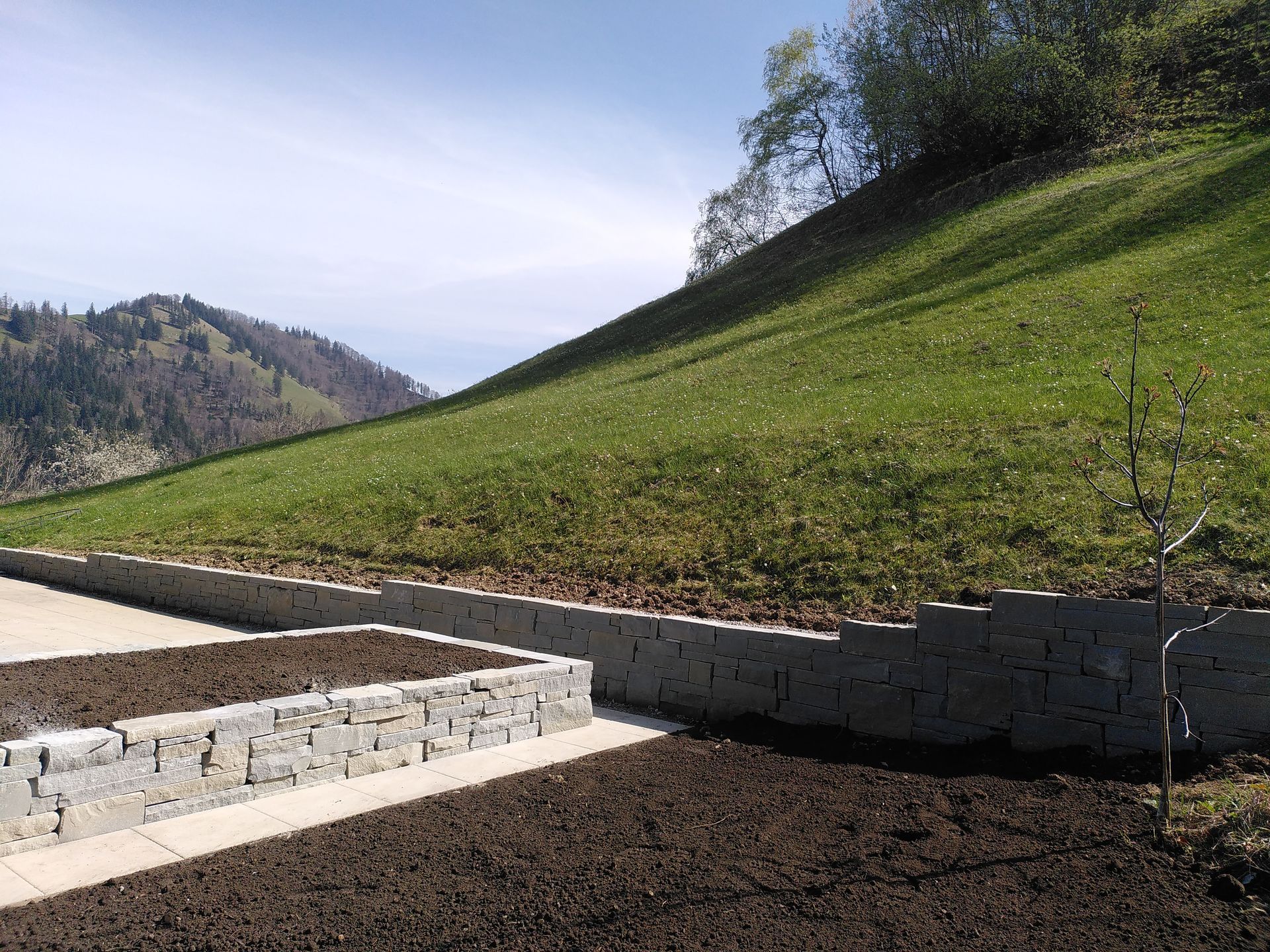 Sonnenbeschienener, grasbewachsener Hang mit Steinterrassen und einem bewaldeten Tal in der Ferne