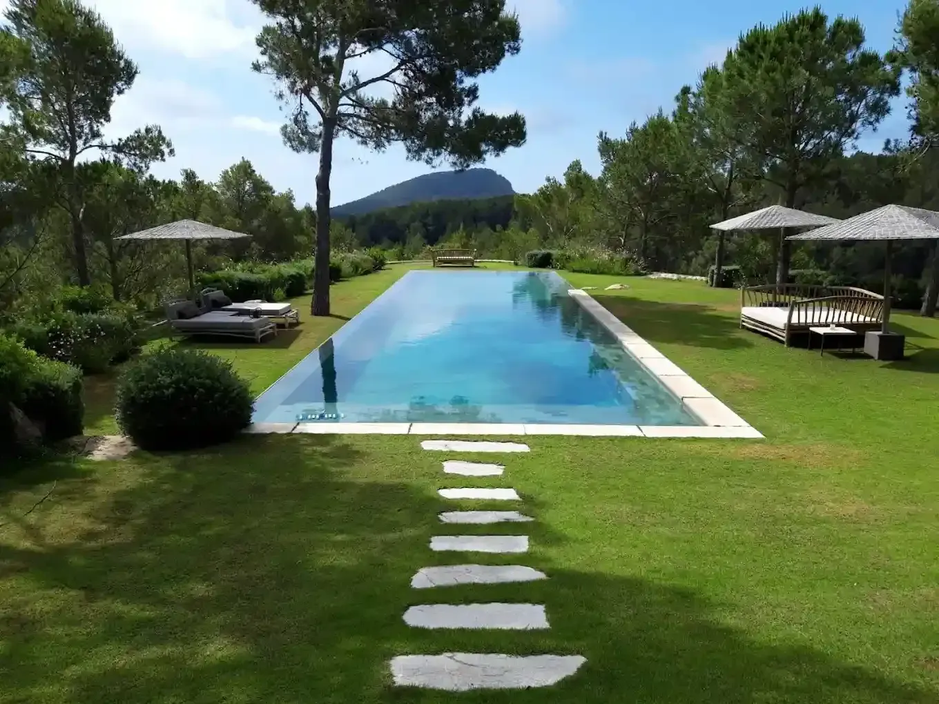 Piscina rectangular alargada con escalones sobre césped, flanqueada por árboles y zonas para sentarse.