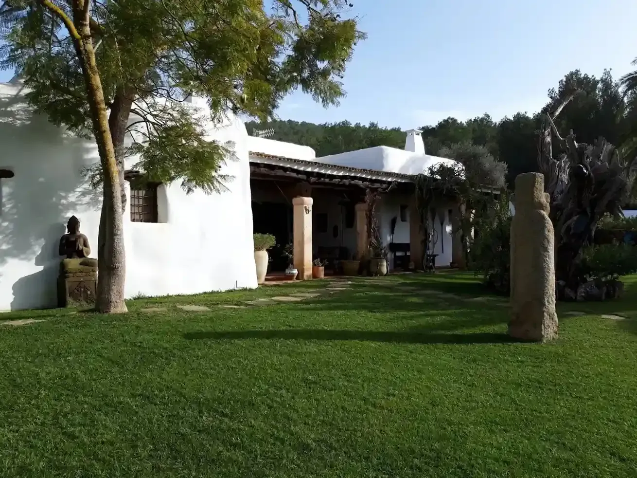 Edificio encalado con un porche cubierto y un patio con césped; una escultura y un árbol están en primer plano.