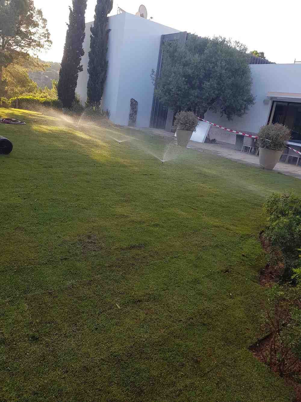 Aspersores de césped riegan un césped verde frente a un edificio blanco con árboles.