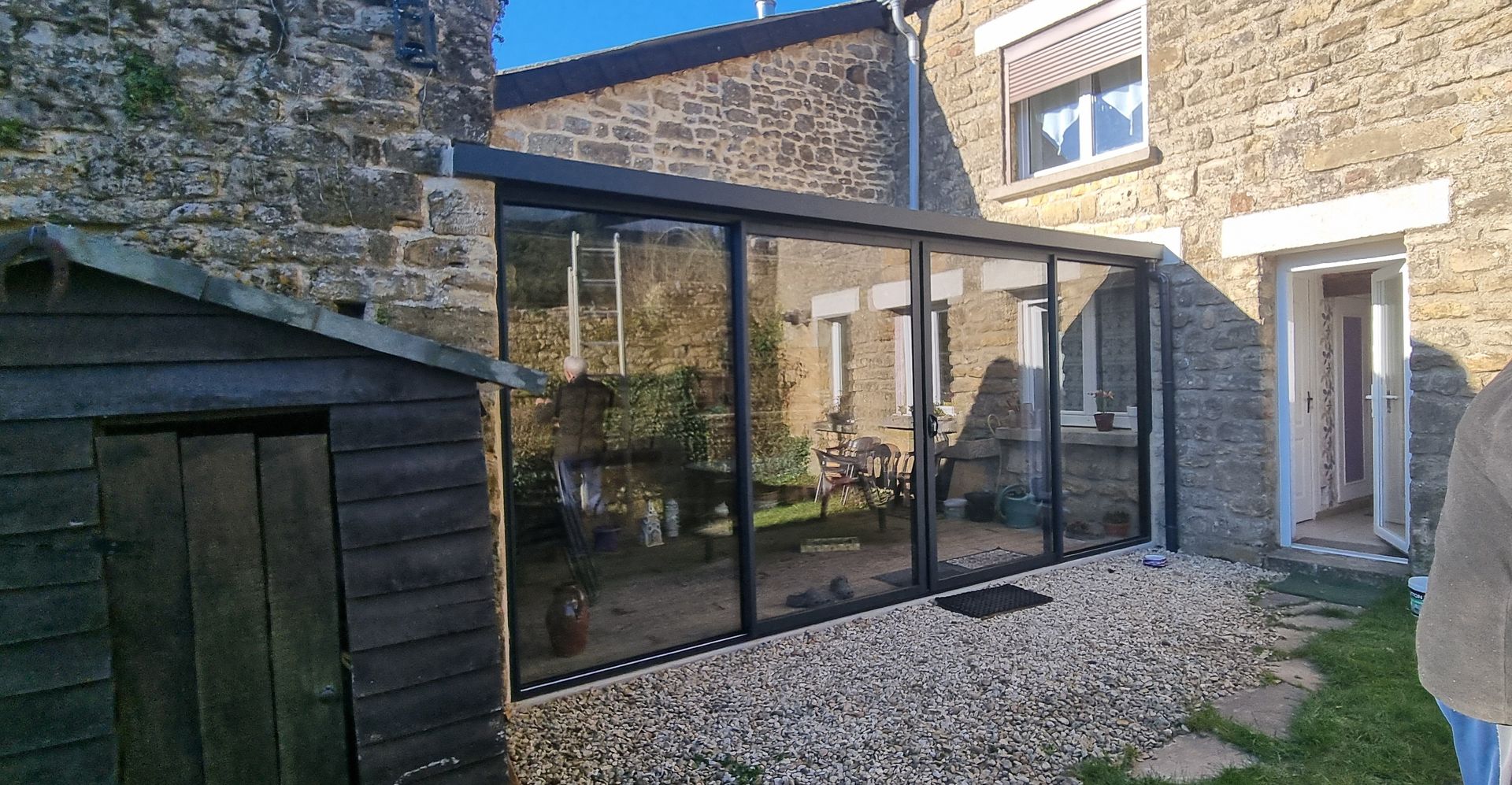 Véranda vitrée entre des bâtiments en pierre, porte ouverte, sol en gravier, plantes visibles, journée ensoleillée.