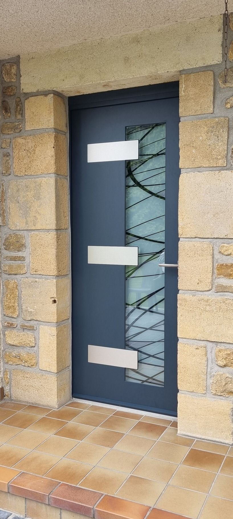 Porte d'entrée bleue avec panneau de verre et accents métalliques horizontaux, encastrée dans une allée en pierre.