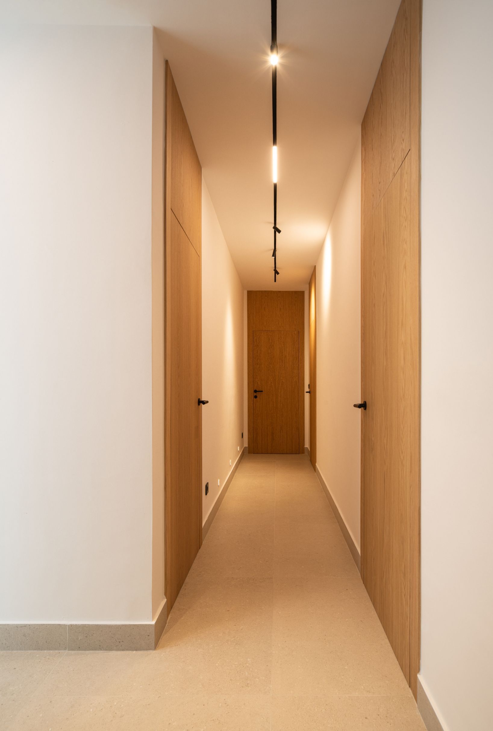 Un pasillo largo y estrecho con paredes blancas, puertas de madera clara, suelo beige y una lámpara de techo lineal