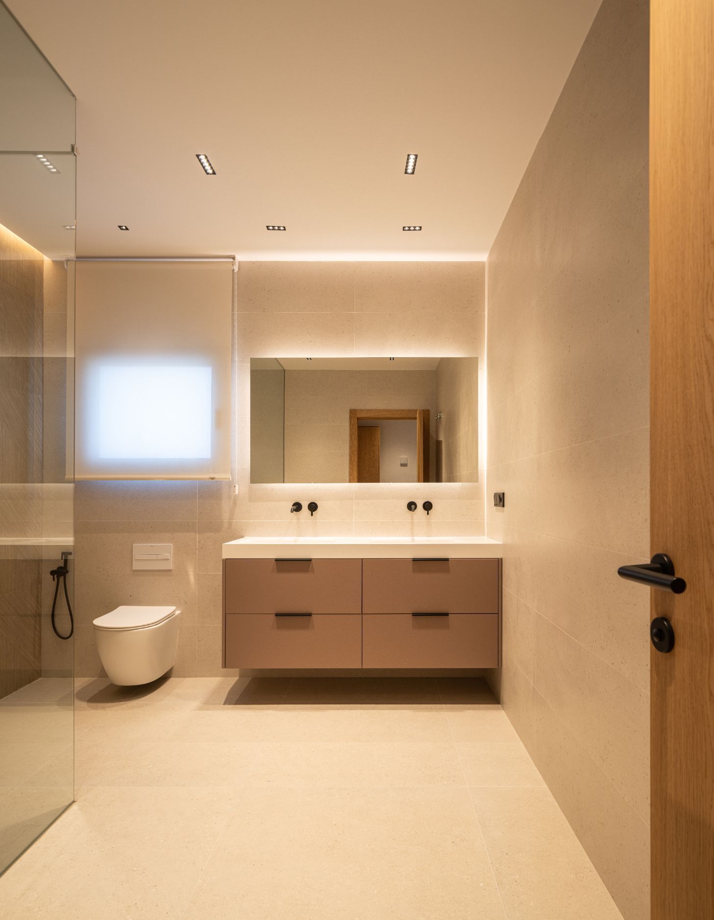 Baño moderno con puerta de madera clara, paredes alicatadas en color beige, tocador flotante con dos lavabos