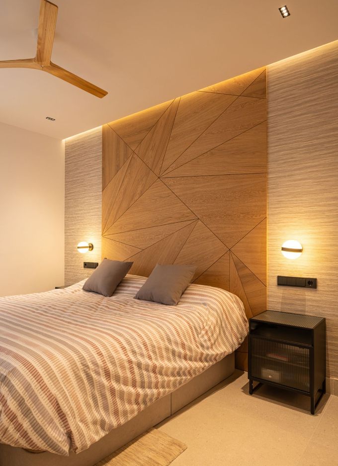 Un dormitorio con cabecero de madera con paneles geométricos, iluminación ambiental cálida y una cama con una colcha a rayas.