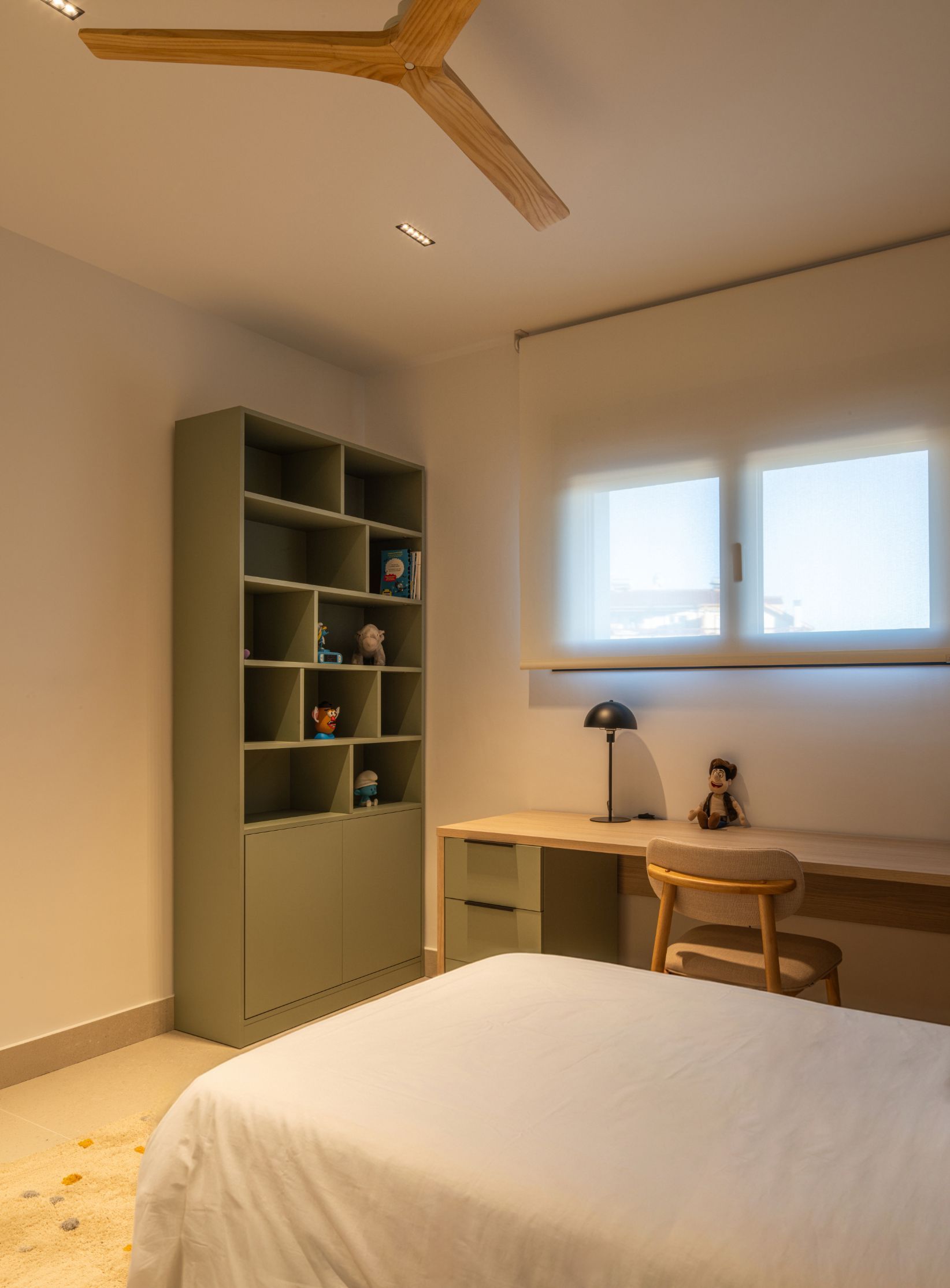 Un dormitorio moderno con cama, estantería verde, escritorio con lámpara y silla, y ventilador de techo de madera.