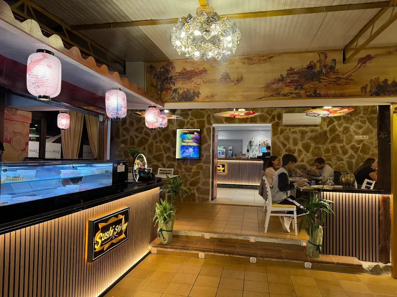 Interior de un restaurante japonés con linternas decorativas, una barra de sushi y clientes cenando.