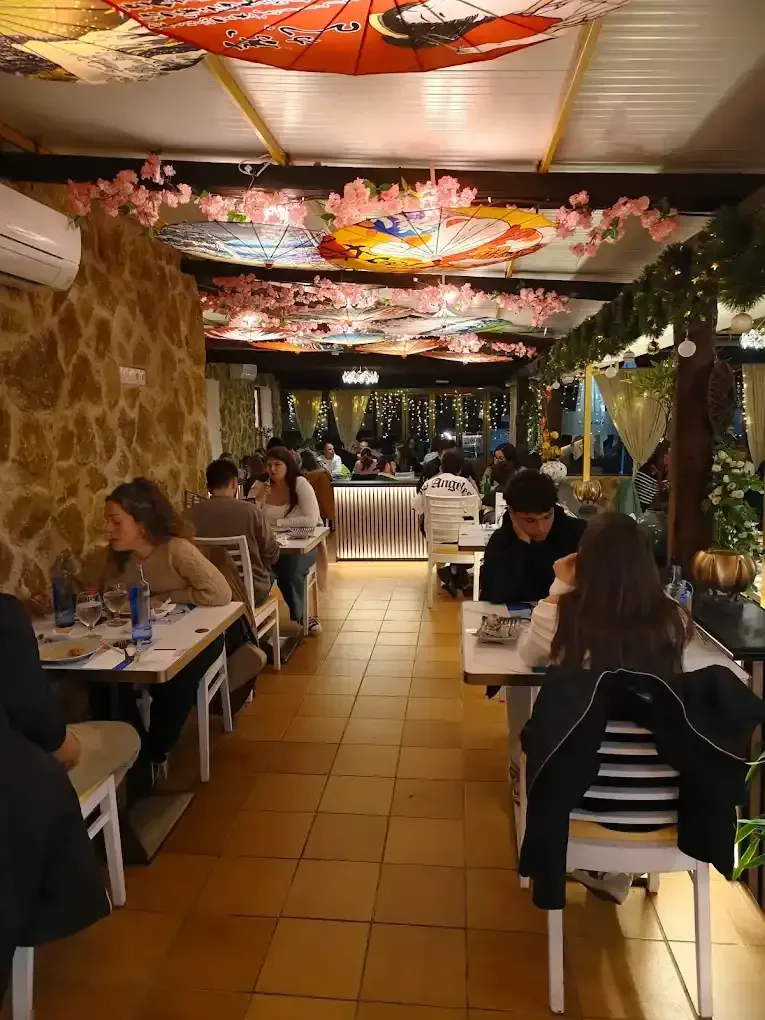 Interior del restaurante, comensales en las mesas. Flores de cerezo y sombrillas japonesas decoran el techo. Muro de piedra a la izquierda.