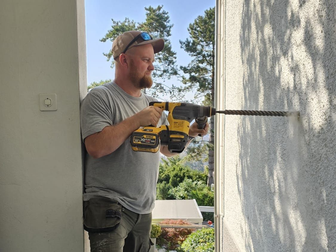 Ein Mann bohrt in eine Stuckwand im Freien. Er trägt eine Mütze, einen Gehörschutz und ein graues Hemd.