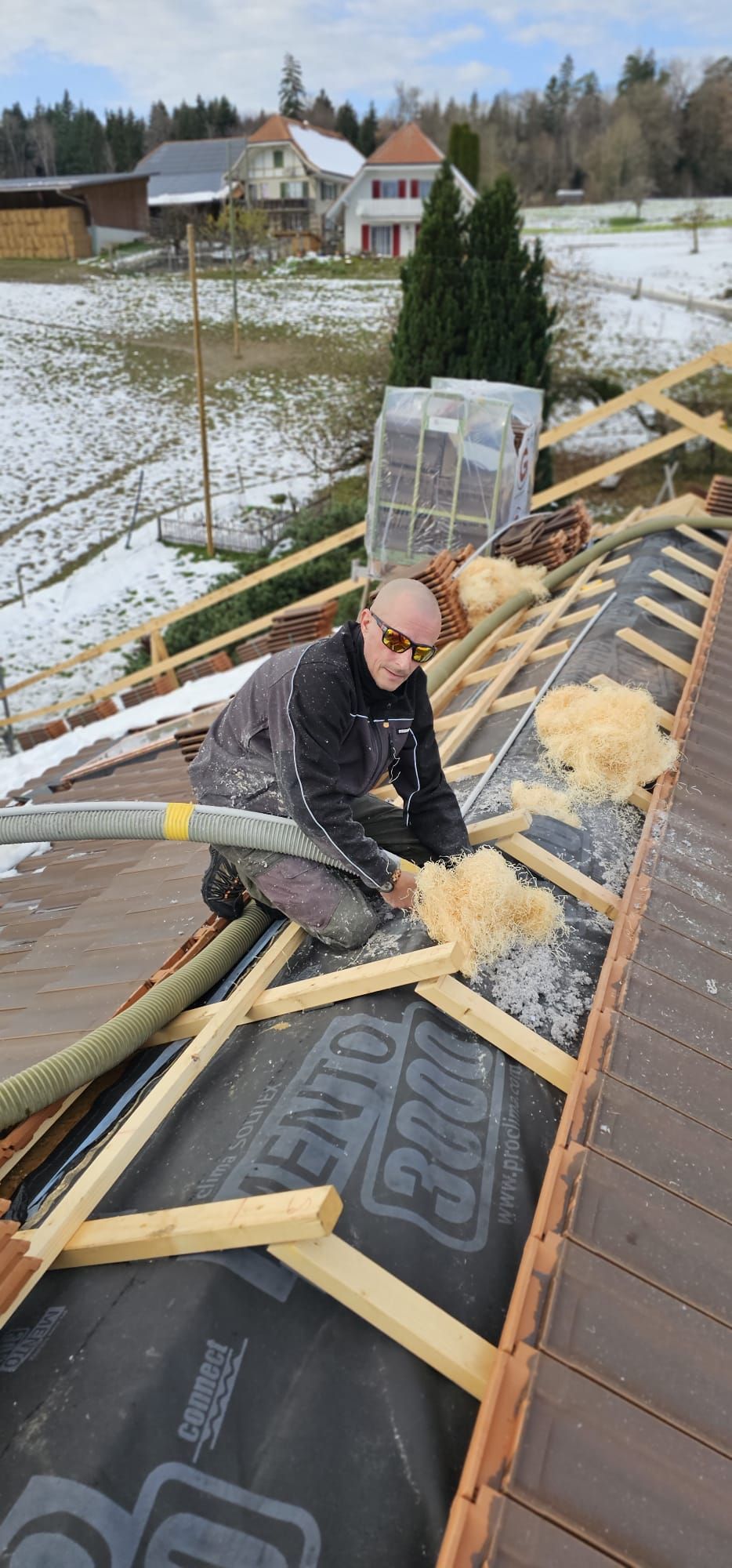 Ein Dachdecker mit Sonnenbrille kniet auf einem Dach und isoliert. Im Hintergrund sind verschneite Häuser zu sehen.