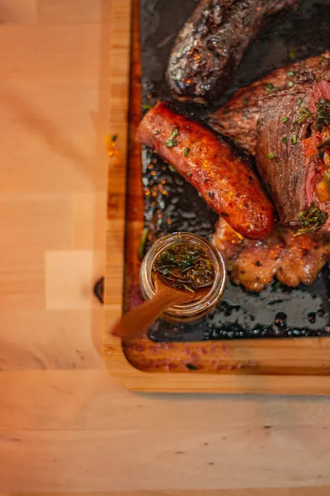 Salchichas y carne a la parrilla sobre una tabla de madera con un pequeño frasco de condimento.