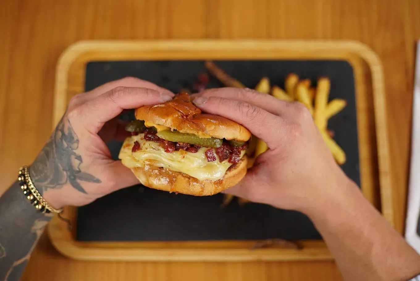 Manos sosteniendo una hamburguesa con queso sobre una bandeja con papas fritas en una mesa de madera.