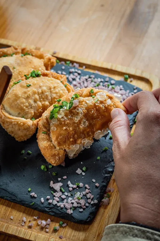 Una mano sostiene un pastel frito en una bandeja con dos pasteles, espolvoreados con sal gruesa y hierbas.