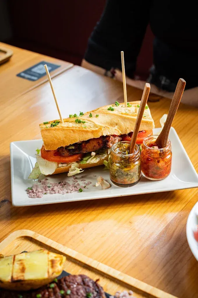 Un sándwich en un plato blanco con dos frascos de salsa sobre una mesa de madera.