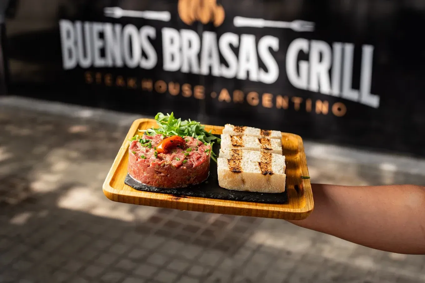 Mano sosteniendo un plato de bistec a la parrilla y verduras frente al letrero de Buenos Brasas Grill.