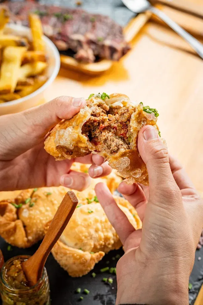 Unas manos abren un panecillo relleno de carne y hierbas sobre una mesa con patatas fritas y salsas.