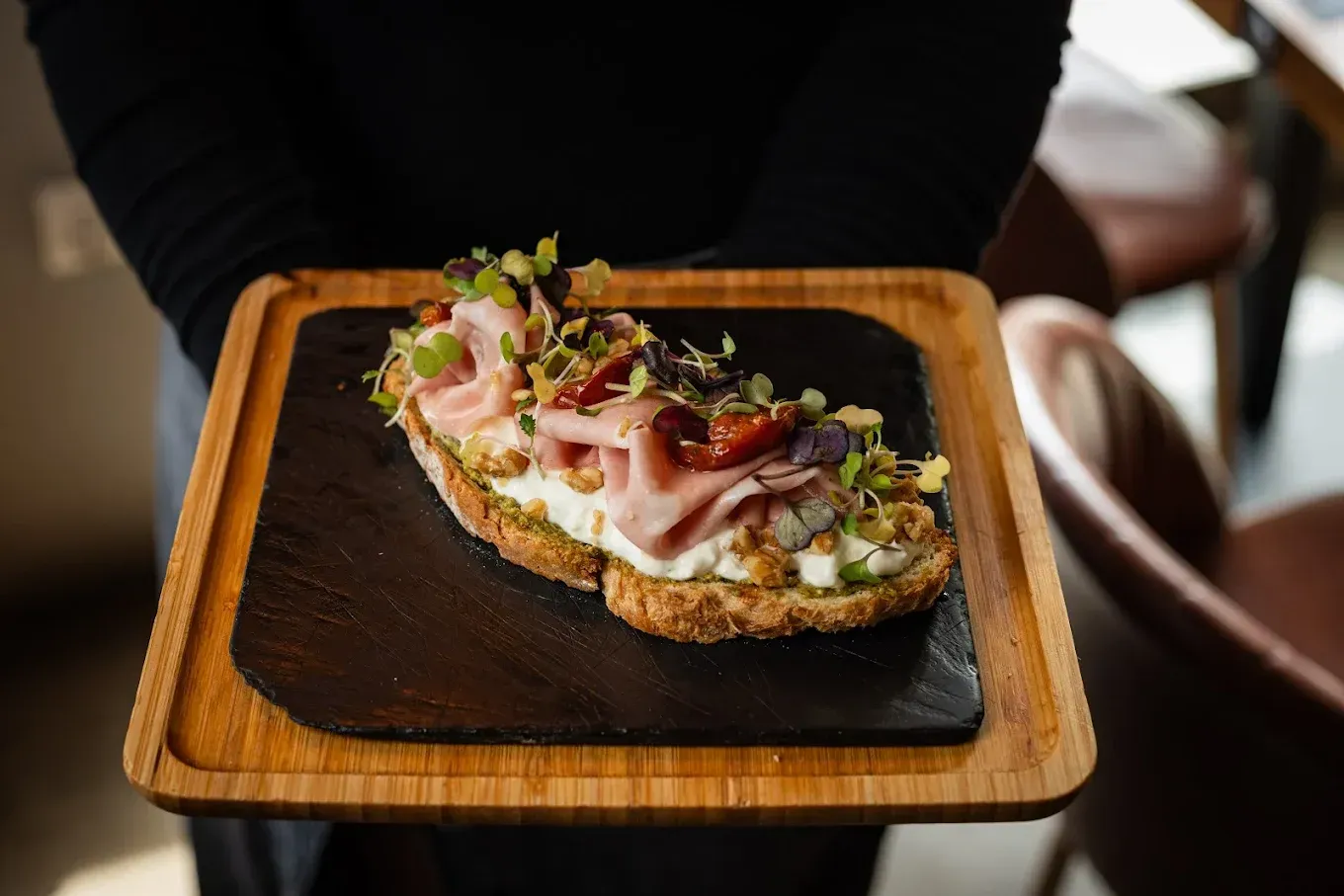 Tostada con ingredientes sobre un plato de madera sostenido por una persona con una camisa oscura.