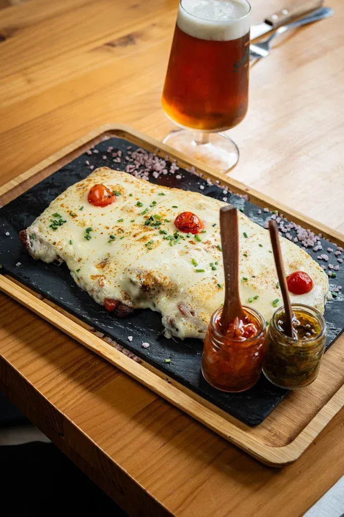 Plato gratinado con queso y tomates cherry en una bandeja de madera, servido con cerveza y dos salsas.