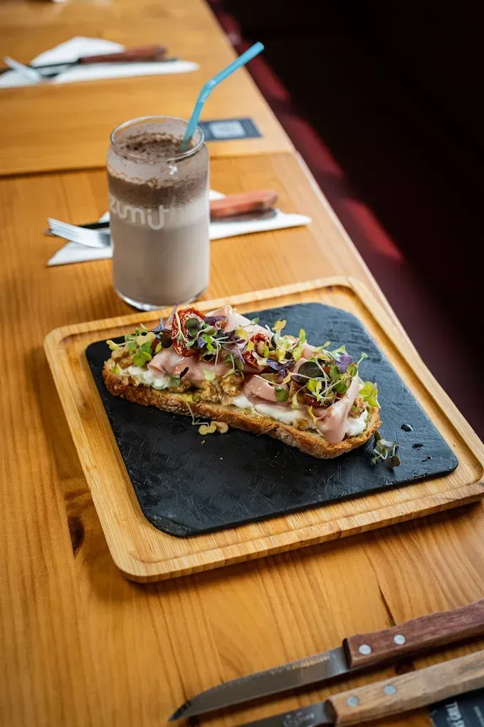 Un sándwich sobre una tabla de pizarra negra con una bebida helada sobre una mesa de madera y palillos chinos al lado.