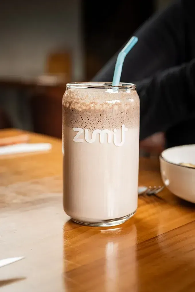 Vaso alto de leche con chocolate y pajita azul sobre una mesa de madera, con el logotipo de “zumu”.