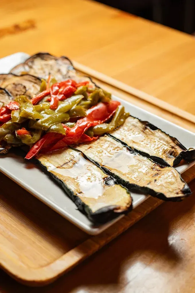 Berenjena a la parrilla emplatada con pimientos rojos y verdes sobre una mesa de madera.