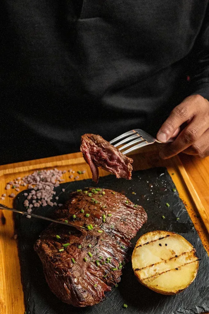 Persona sirviendo filete en rodajas con un tenedor junto a una patata asada en una tabla de madera.