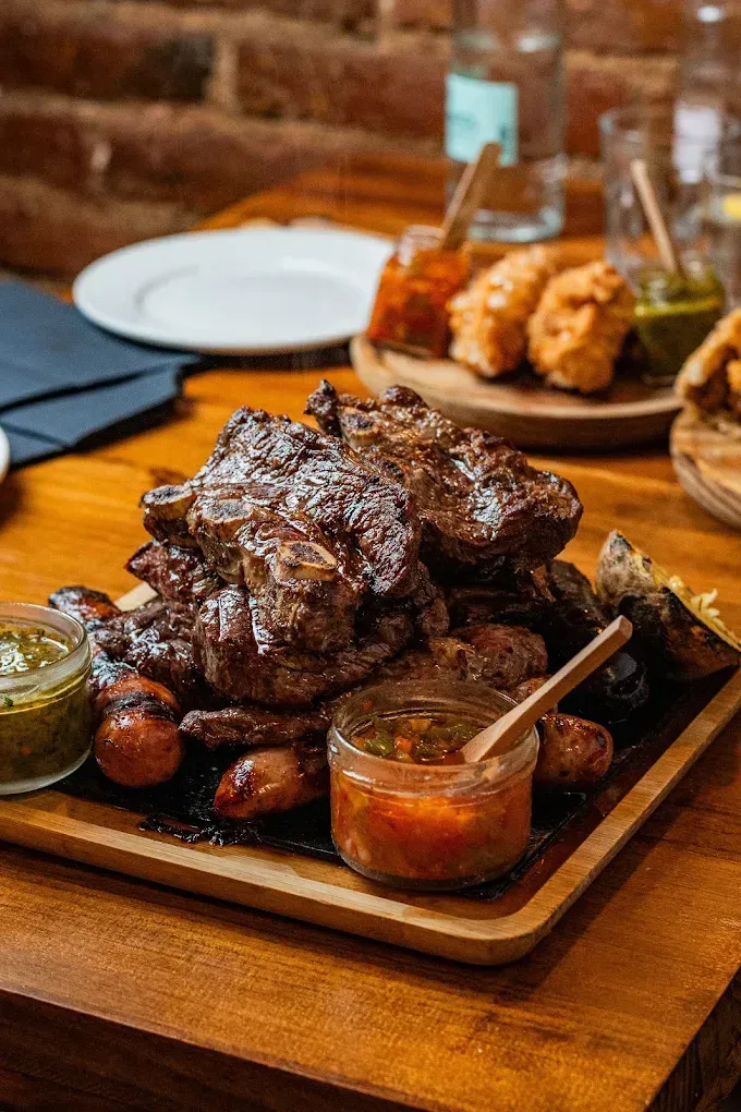 Plato de carne a la parrilla con salsas para mojar sobre una mesa de madera en un ambiente de restaurante rústico.