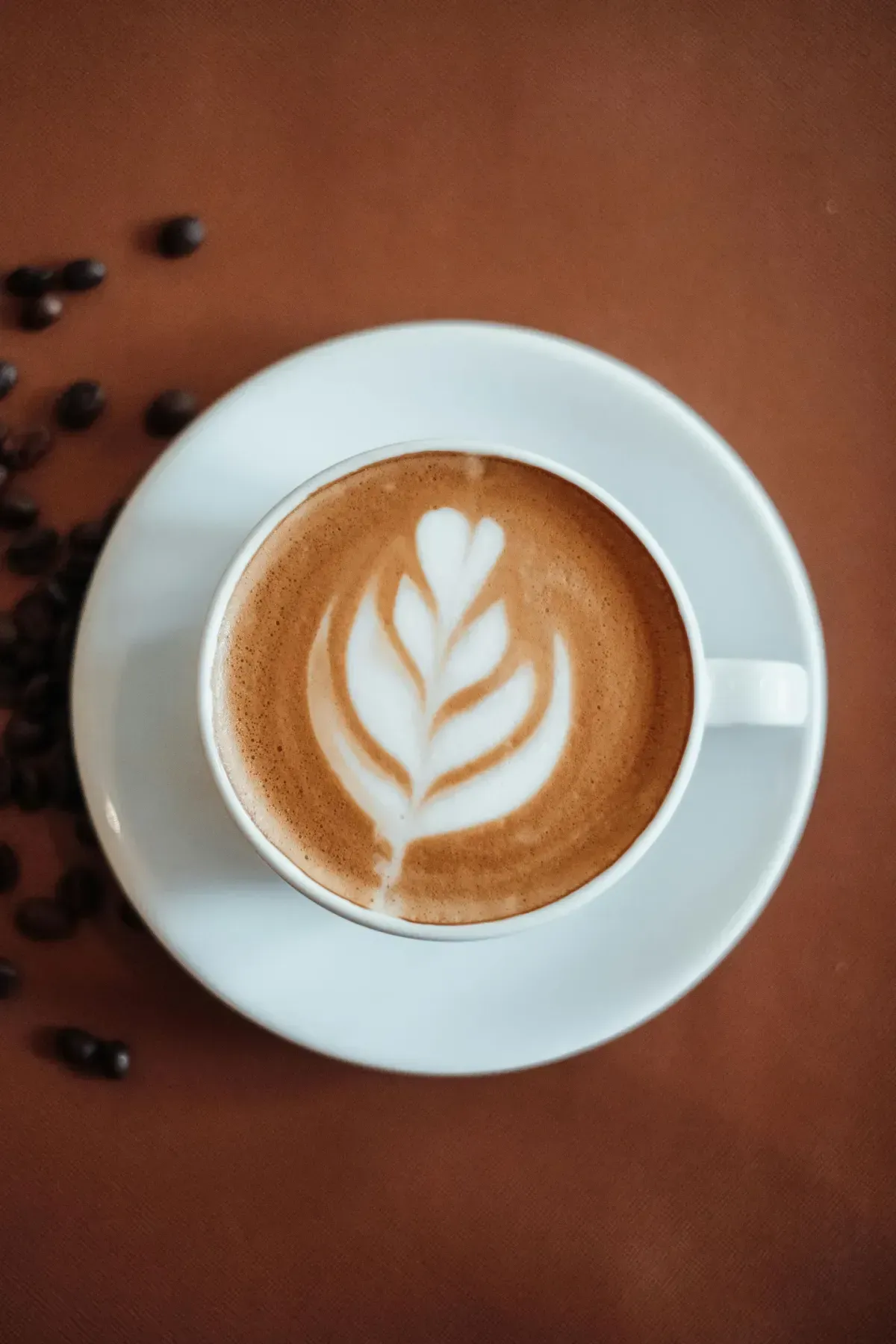 Vista superior de un café con leche decorado con espuma de hojas en una taza blanca sobre una mesa marrón con granos de café.