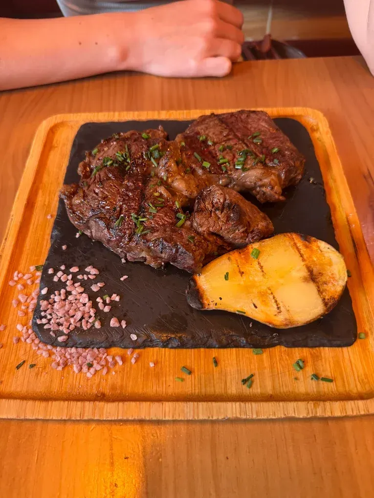 Filete a la parrilla y patata asada sobre una tabla de madera, con una mano apoyada al fondo.