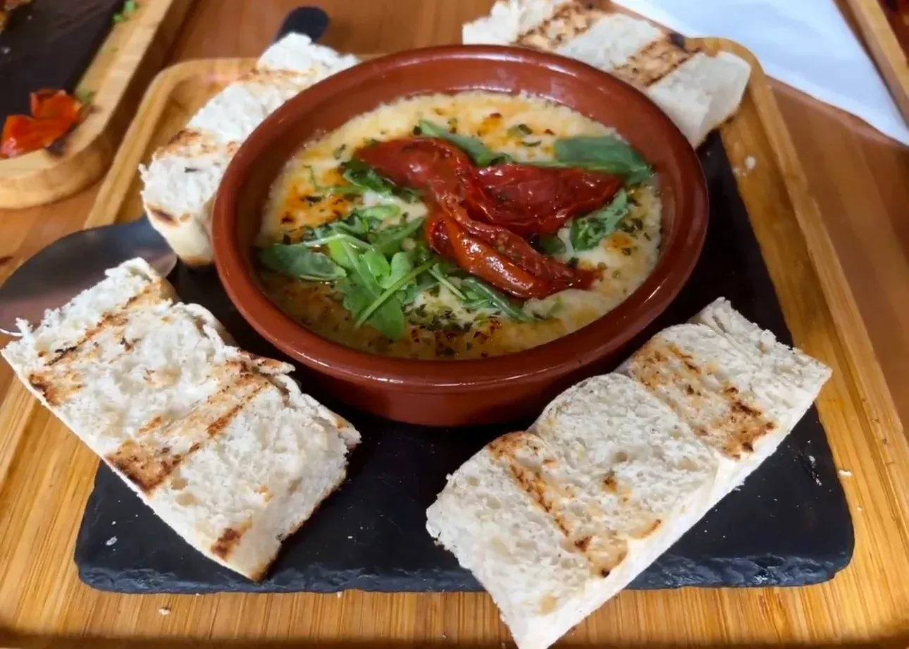 Salsa picante en un cuenco de barro con hierbas y pimientos rojos, acompañada de pan tostado sobre una tabla de madera.