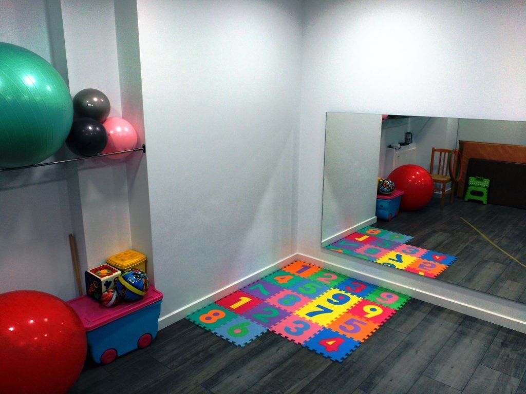 Una habitación con pelotas de ejercicio, un espejo y coloridas colchonetas de espuma sobre un suelo gris.