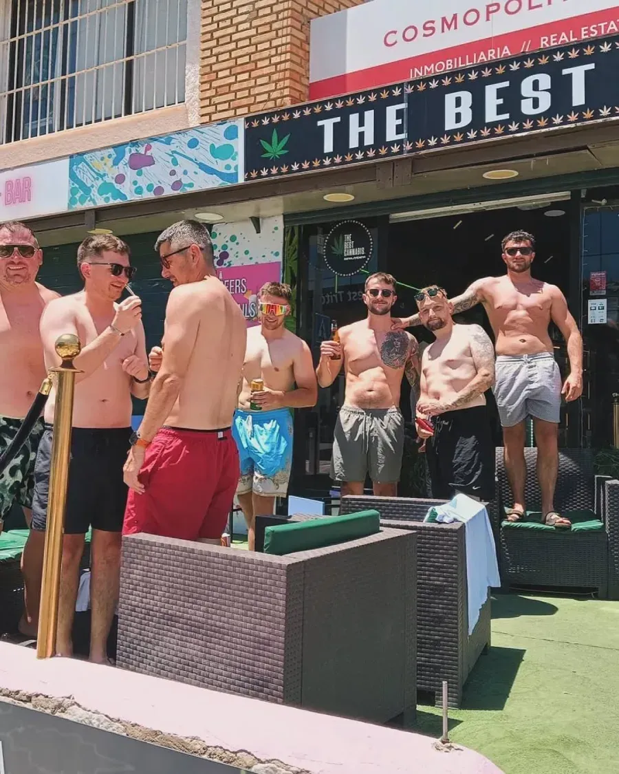 Grupo de hombres sin camisa afuera de un bar, algunos con bebidas. El cartel del bar dice 